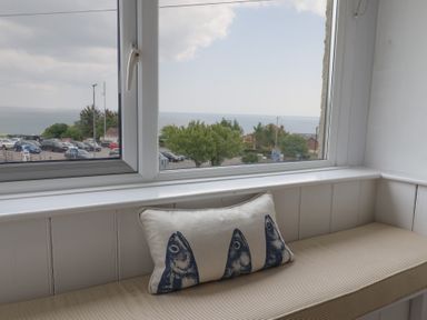 A window seat with a cushion featuring three fish heads and a view of parked cars trees and the sea at Ancient Lights in Lyme Regis