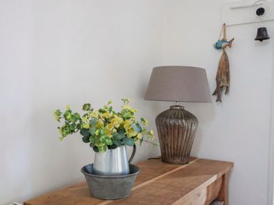 A wooden table with a metal pitcher holding green and yellow flowers next to a lamp with a brown shade and a decorative fish hanging on the wall at Ancient Lights in Lyme Regis