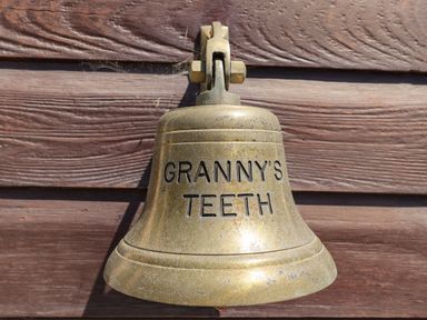 A brass bell mounted on a wooden wall with the words Granny's Teeth engraved on it at Grannys Teeth in Lyme Regis