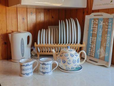 A kitchen counter with a white electric kettle two mugs labeled skipper and crew a teapot with nautical designs a dish rack with plates and a tray with text at Granny's Teeth Lyme Regis