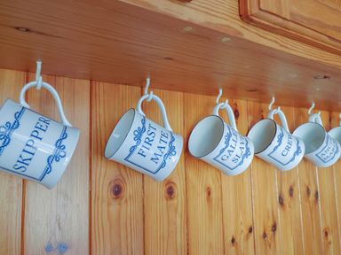 A row of ceramic mugs labeled skipper first mate galley slave crew and anchor man hanging on hooks on a wooden wall