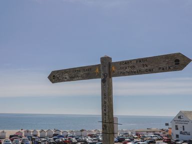 A wooden signpost with directions for coast paths with parked cars and beach huts near the sea at Granny's Teeth in Lyme Regis