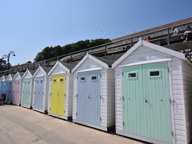 A row of small beach huts with pastel-colored doors along a promenade with people sitting on benches above at Granny's Teeth in Lyme Regis