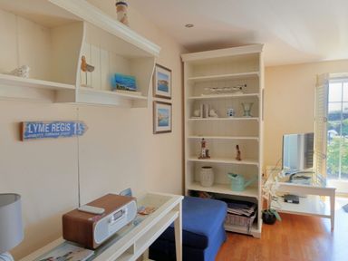 A living room with shelves holding decorative items a television on a white table and a blue ottoman at 2 Buckfields Lyme Regis