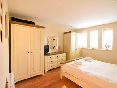 A bedroom with a bed with white linens two wardrobes a dresser with a mirror and a television near three windows at 2 Buckfields Lyme Regis