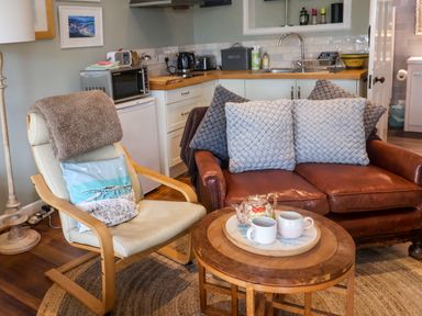 A sitting area with a leather sofa and cushioned chair next to a small wooden table with a teapot and cups in a room with a kitchenette at The Apartment at Queen Anne's Lodge Lyme Regis