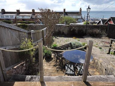 A backyard with a wooden deck, a round glass table, benches, plants, and a view of nearby houses and the sea at 10 Cobb Terrace in Lyme Regis