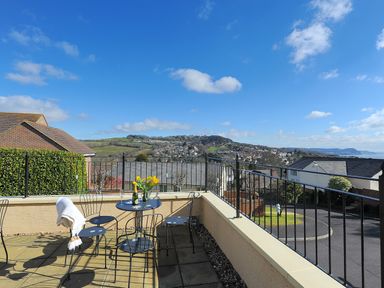 A balcony with a table and chairs overlooking a residential area and hills at Jurassic Sea View Lyme Regis
