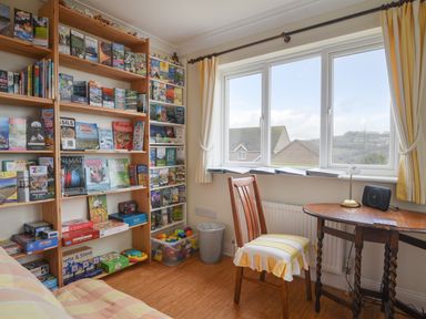 A room with bookshelves filled with books and games a chair and a wooden table under a window with yellow curtains at Jurassic Sea View in Lyme Regis