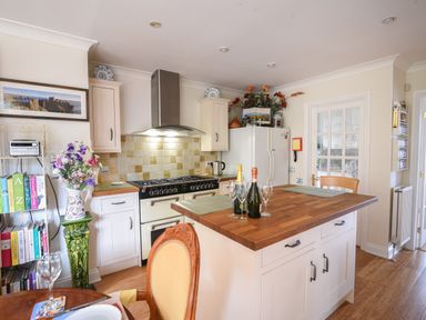 A kitchen with a wooden island countertop with bottles and glasses a stove a white refrigerator a door a bookshelf and a vase with flowers at Jurassic Sea View in Lyme Regis