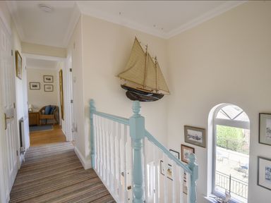 A hallway with a striped carpet and white and blue banister with a large model sailboat on the wall and an arched window with framed pictures near Jurassic Sea View Lyme Regis
