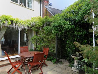 An outdoor patio with a wooden table and four chairs surrounded by greenery and a birdbath