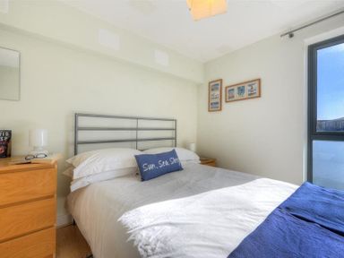 A bedroom with a bed white bedding and blue pillow beside wooden bedside tables with lamps and framed pictures on the wall at 8 Driftwood in West Bay