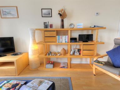A living room with a wooden shelving unit holding books vases and a clock next to a lamp and a television at 8 Driftwood in West Bay
