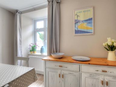 A dining area with a table covered with a polka dot cloth a window with curtains a sideboard with dishes and flowers and a Lyme Regis poster on the wall at Mermaid House in Lyme Regis