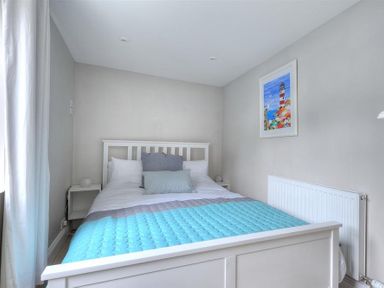 A bedroom with a white bed frame a bed with pillows a blue quilted blanket a white nightstand a colorful framed picture on the wall and a radiator at Mermaid House in Lyme Regis