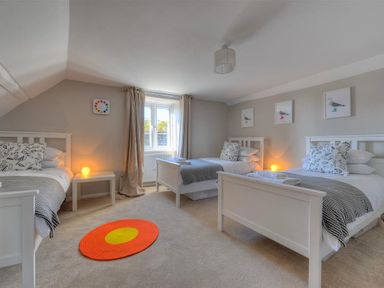 A bedroom with three single beds side by side with patterned pillows and grey throws with a window and small night tables with lamps at Mermaid House in Lyme Regis