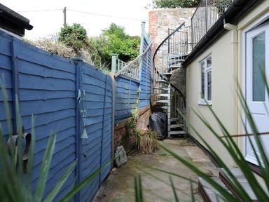 A narrow outdoor area with blue wooden fencing spiral metal staircase and cream wall with windows at Mermaid House in Lyme Regis
