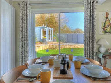 A dining table set with bowls cups and cutlery in front of sliding glass doors leading to a garden with a shed at Woody View in Colyton