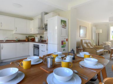 A kitchen and dining area with white cabinets yellow mugs and white bowls on a wooden table and a beige living room with leather chairs at Woody View in Colyton