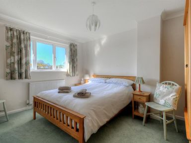 A bedroom with a wooden double bed with white bedding two bedside tables with lamps a window with curtains a wooden wardrobe and a chair with a cushion at Woody View in Colyton