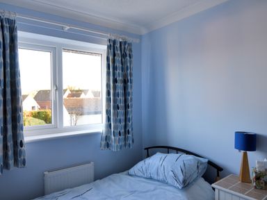 A bedroom with a single bed beside a window with patterned curtains and a bedside table with a blue lamp at Woody View in Colyton