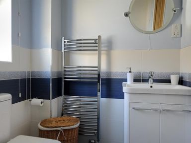 A bathroom with a white sink and cabinet a round mirror a chrome towel rack a toilet a wicker laundry basket and a tiled wall at Woody View in Colyton