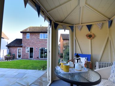Inside a garden gazebo with a glass table holding a tea set and chairs with cushions overlooking a backyard and a two-story brick house at Woody View in Colyton