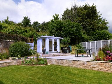 A garden with a small white shed on a stone patio and green lawn at Woody View in Colyton