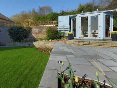 A garden with a lawn patio and a small blue wooden summerhouse at Woody View in Colyton