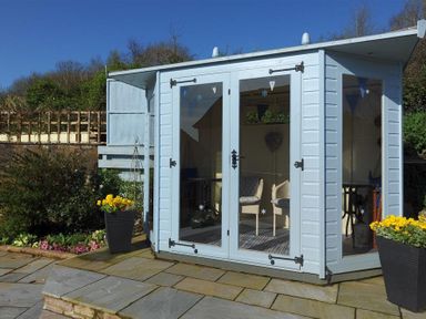 A small garden summerhouse with glass doors and chairs inside surrounded by flowers and a stone patio at Woody View in Colyton