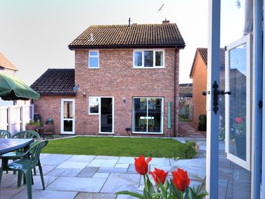 Backyard with patio, green lawn, red flowers, outdoor table with chairs, and two-story brick house with windows and doors at Woody View Colyton