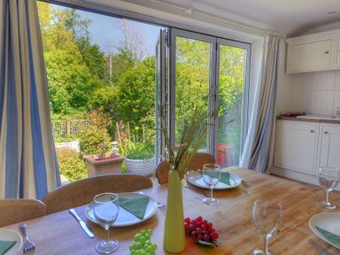 A dining table set with plates glasses and cutlery beside open glass doors overlooking a garden at 24 Mill Green Lyme Regis