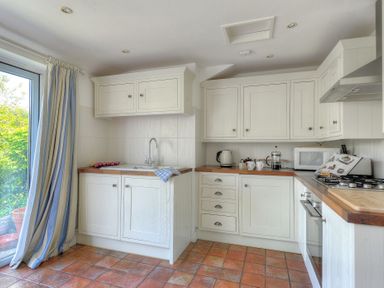 A kitchen with white cabinets wooden countertops a sink a kettle a coffee press a microwave and a stove next to a sliding glass door with curtains at 24 Mill Green in Lyme Regis