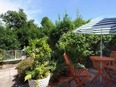 A patio with wooden chairs and table with a striped umbrella surrounded by plants and trees at 24 Mill Green in Lyme Regis