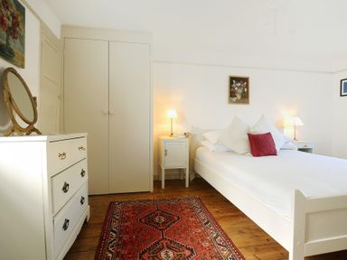 A bedroom with a white bed side tables lamps a dresser mirror wardrobe and a red patterned rug at 24 Mill Green Lyme Regis