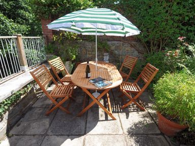 An outdoor patio with a wooden table and six chairs under a striped umbrella with plants and greenery around at 24 Mill Green in Lyme Regis