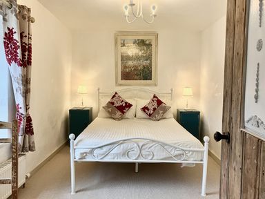 A bedroom with a white metal bed frame two bedside tables with lamps pillows on the bed a painting on the wall and floral curtains at 24 Mill Green in Lyme Regis