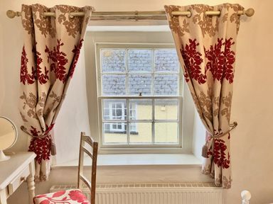 A window with patterned curtains beside a wooden chair and a small white table with a mirror at 24 Mill Green in Lyme Regis