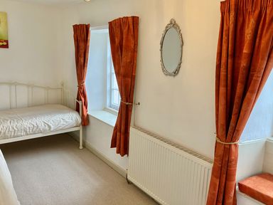 A bedroom with a single bed beside a window with orange curtains an oval mirror on the wall radiator below the mirror and a window seat with an orange cushion at 24 Mill Green in Lyme Regis