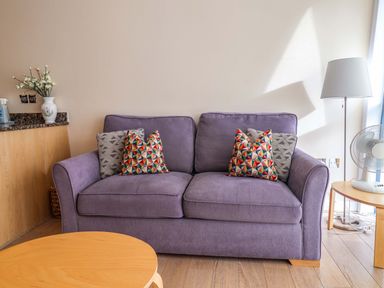 A purple sofa with patterned cushions in a room with wooden tables and a floor lamp at 2 Driftwood in West Bay