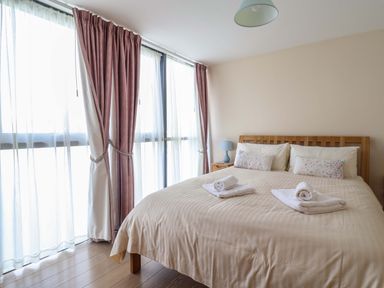A bedroom with a wooden bed with beige bedding and two folded towels on it a bedside table with a blue lamp and large windows with sheer and pink curtains at 2 Driftwood in West Bay