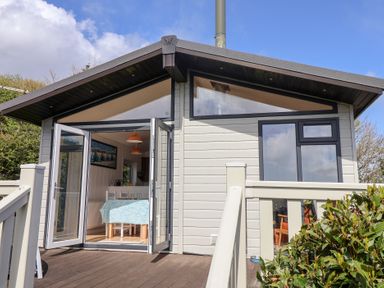 A small lodge with an open door leading to a dining area with a table and chairs on a wooden deck at High Bullen Lodge in Morcombelake