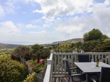 A balcony with a table and chairs overlooking a garden and countryside with a view of the sea at High Bullen Lodge in Morcombelake