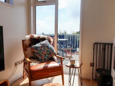 A leather armchair with a pillow and fur cover next to a small wooden side table with a cup by a glass door opening to a balcony at High Bullen Lodge in Morcombelake