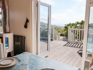 A dining area with a table set with plates and glasses next to a door opening to a wooden deck with railings and plants outside at High Bullen Lodge in Morcombelake