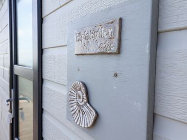 A gray wooden plaque with a shell decoration and the text High Bullen Lodge mounted on an exterior wall next to a door at High Bullen Lodge in Morcombelake