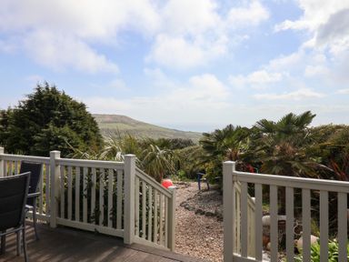 A wooden deck with chairs overlooking a garden path with palm trees and hills in the distance at High Bullen Lodge in Morcombelake