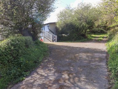 An outdoor area with a pathway leading to a white wooden building surrounded by bushes and trees at High Bullen Lodge in Morcombelake