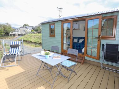 An outdoor deck with a small table and two chairs and a striped lounge chair next to a green cabin with open double doors showing a living area at Llety'r Bugail 1 in Llanrug
